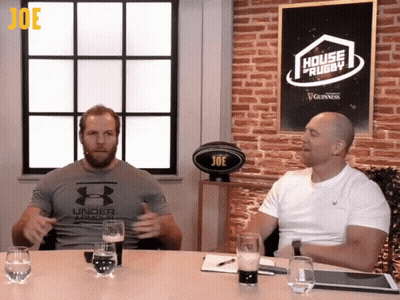 Two Men Sitting At A Table With A Sign That Says House Of Rugby GIF