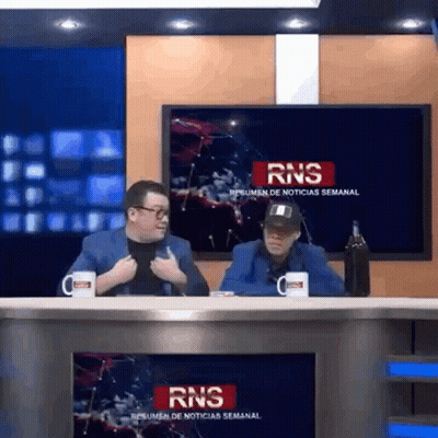 Two Men Sitting At Table In Front Of RNS Sign GIF
