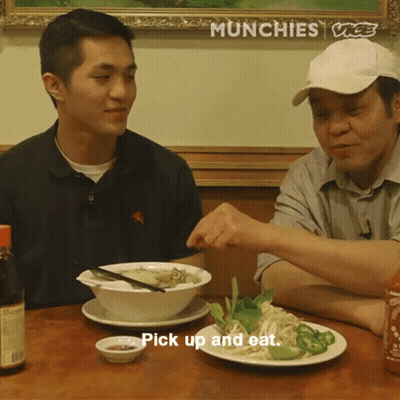Two Men Sitting At Table With Bowl Of Food Saying Pick Up And Eat GIF