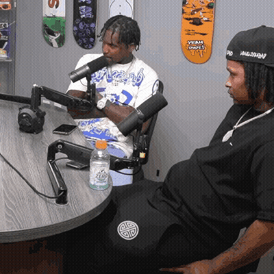 Two Men Sitting At Table With Microphone And Skateboards GIF