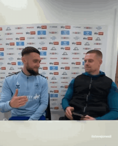 Two Men Sitting In Front Of Sky Bet Wall GIF