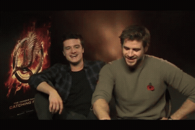 Two Men Sitting Next To Each Other In Front Of A Hunger Games Poster GIF