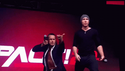 Two Men Stand In Front Of A Pac Wrestling Logo GIF