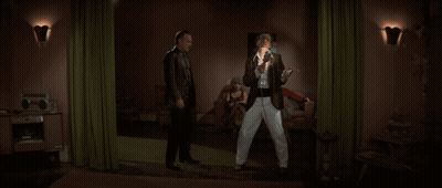 Two Men Standing In A Living Room With A Green Curtain GIF