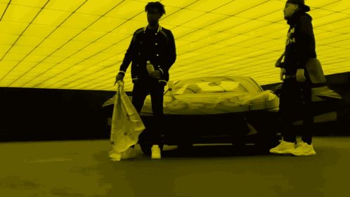 Two Men Standing In Front Of A Car With One Wearing A Black Jacket With The Letter M On The Front GIF