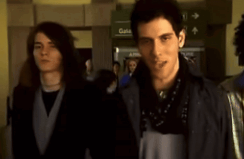 Two Men Standing In Front Of A Green Sign That Says Gates GIF