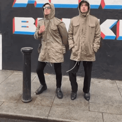 Two Men Standing On A Sidewalk In Front Of A Wall That Has The Letter B Painted On It GIF