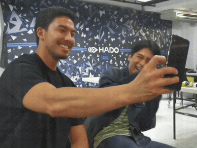 Two Men Take A Selfie In Front Of A Wall That Says Hado Blue GIF