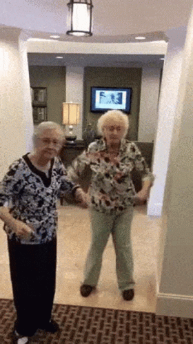Two Older Women Are Dancing In A Hallway In Front Of A Tv GIF