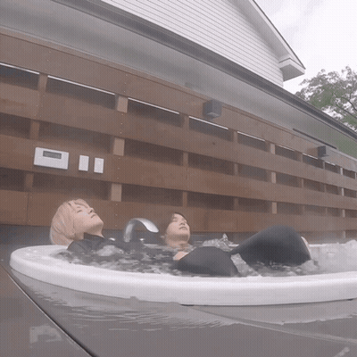Two People Are Laying In A Hot Tub Outside A House With A Clock On The Wall GIF
