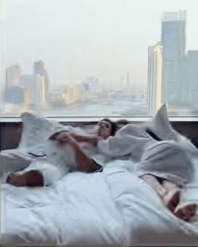 Two People Are Laying On A Bed In Front Of A Window With A City In The Background GIF