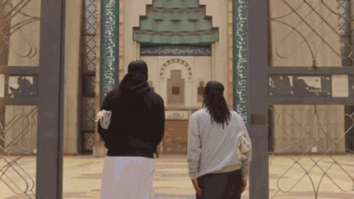 Two People Are Standing In Front Of A Building With Arabic Writing On The Wall GIF