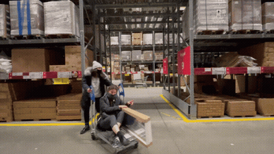 Two People Pushing A Cart In A Warehouse With A Red Sign That Says Ikea GIF