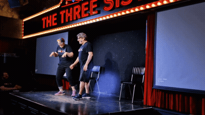 Two People Stand On A Stage Under A Neon Sign That Says The Three Sisters GIF