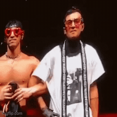 Two Shirtless Men Are Standing Next To Each Other On A Stage . One Of The Men Is Wearing Sunglasses And A Scarf . GIF