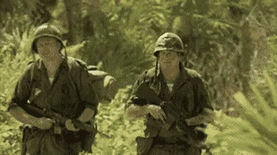 Two Soldiers Are Walking Through A Lush Green Forest . GIF