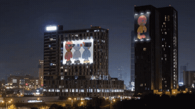 Two Tall Buildings Are Lit Up At Night And One Has A Picture Of Two Cartoon Characters Projected On It GIF