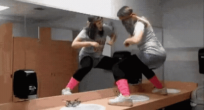 Two Women Are Dancing In Front Of A Mirror In A Public Restroom . GIF