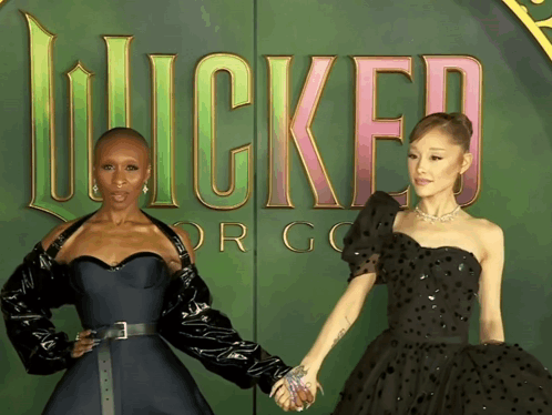 Two Women Are Holding Hands In Front Of A Sign That Says Wicked GIF