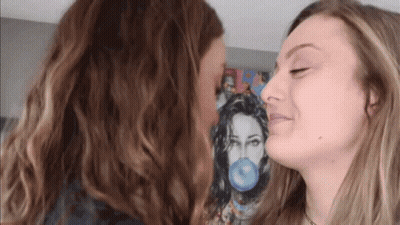 Two Women Are Kissing In Front Of A Painting Of A Woman Blowing A Blue Bubble Gum . GIF