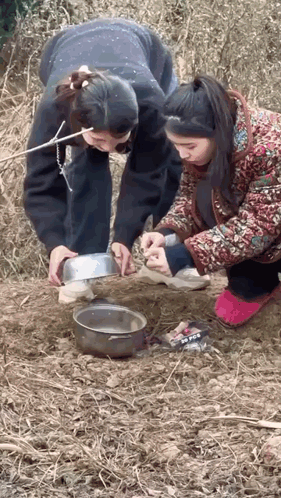 Two Women Are Kneeling Down In The Dirt Looking At A Bowl Of Food . GIF