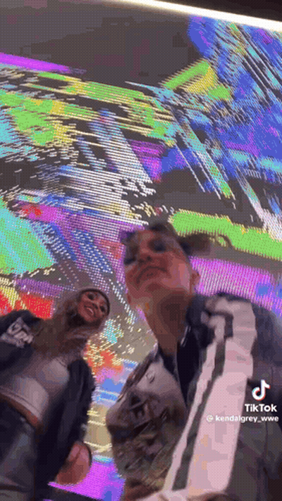 Two Women Are Posing For A Picture In Front Of A Large Screen . GIF