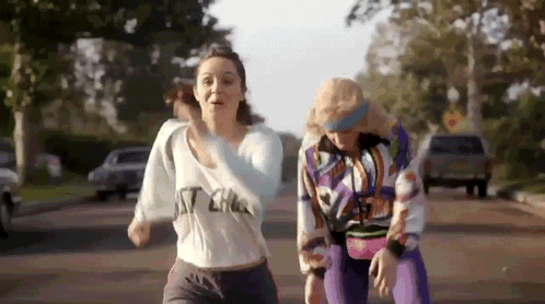 Two Women Are Running Down A Street With One Wearing A Shirt That Says ' I 'M A ' On It . GIF