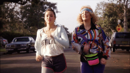 Two Women Are Running Down A Street With One Wearing A Shirt That Says Just Cal GIF