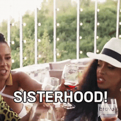 Two Women Are Sitting At A Table Holding Wine Glasses And Toasting With The Words Sisterhood GIF