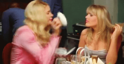 Two Women Are Sitting At A Table Talking To Each Other . One Of The Women Is Holding A Piece Of Paper In Her Hand . GIF
