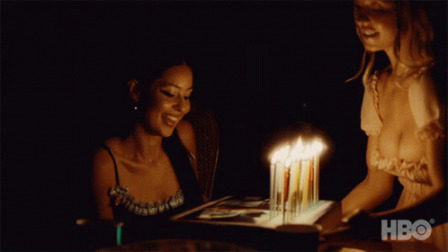 Two Women Are Sitting At A Table With A Cake With Candles On It And Hbo Written On The Bottom GIF
