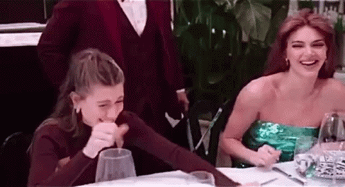 Two Women Are Sitting At A Table With Glasses Of Wine And Laughing . GIF