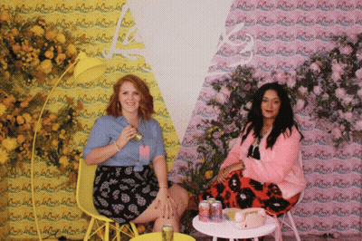 Two Women Are Sitting In Front Of A Wall That Has La Croix Written On It GIF