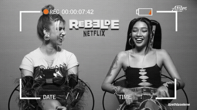 Two Women Are Sitting Next To Each Other In Front Of A Rebelde Netflix Logo GIF