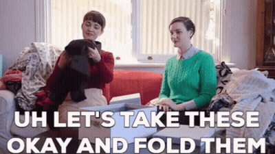 Two Women Are Sitting On A Couch Looking At Clothes And The Caption Says `` Let 'S Take These Okay And Fold Them '' GIF