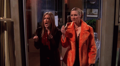 Two Women Are Standing In A Doorway With One Wearing An Orange Coat GIF