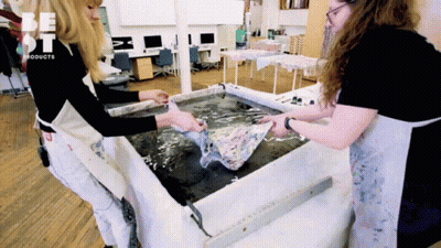 Two Women Are Standing In Front Of A Table That Says Be St Products On It GIF