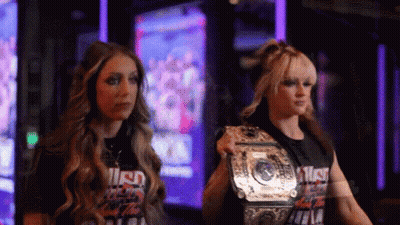 Two Women Are Standing Next To Each Other And One Has A Wrestling Championship Belt GIF