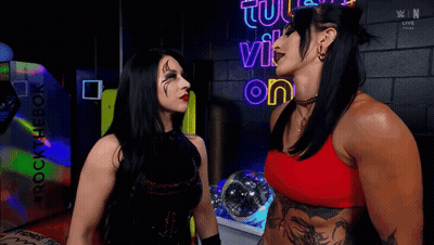Two Women Are Standing Next To Each Other In Front Of A Neon Sign That Says Tu Vi On It . GIF