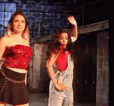 Two Women Are Standing Next To Each Other On A Stage . One Of The Women Is Wearing A Red Top And Overalls . GIF