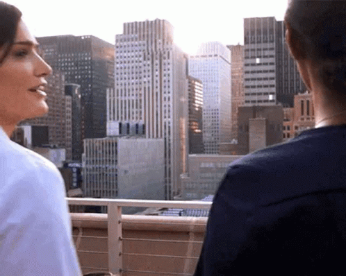 Two Women Are Standing On A Balcony Overlooking A City . GIF