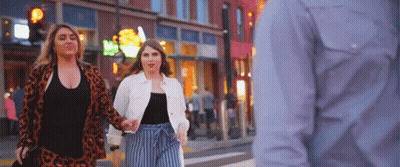 Two Women Are Walking Down A City Street Holding Hands While A Man Looks On . GIF