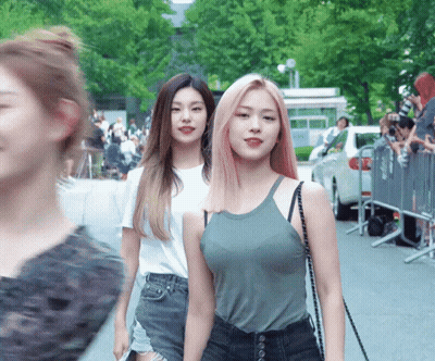 Two Women Are Walking Down A Street With A Crowd Behind Them GIF