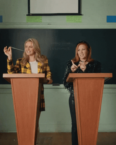Two Women At Podiums With Economics Chalkboard GIF