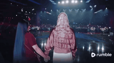 Two Women Holding Hands In Front Of A Stage With A Rumble Logo In The Corner GIF