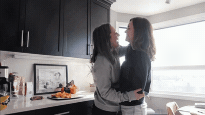 Two Women Hugging In A Kitchen With Black Cabinets And A Picture On The Wall GIF