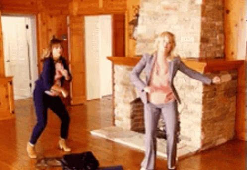 Two Women In Business Suits Are Dancing In A Living Room In Front Of A Fireplace . GIF