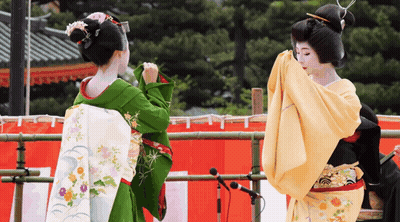 Two Women In Kimonos Are Dancing In Front Of A Red And White Curtain GIF