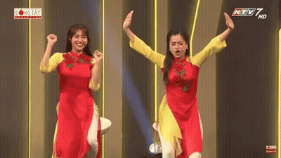 Two Women In Red And Yellow Dresses On Stage With Arms Up GIF