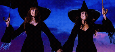 Two Women In Witch Hats Holding Umbrellas In Front Of A Blue Sky GIF